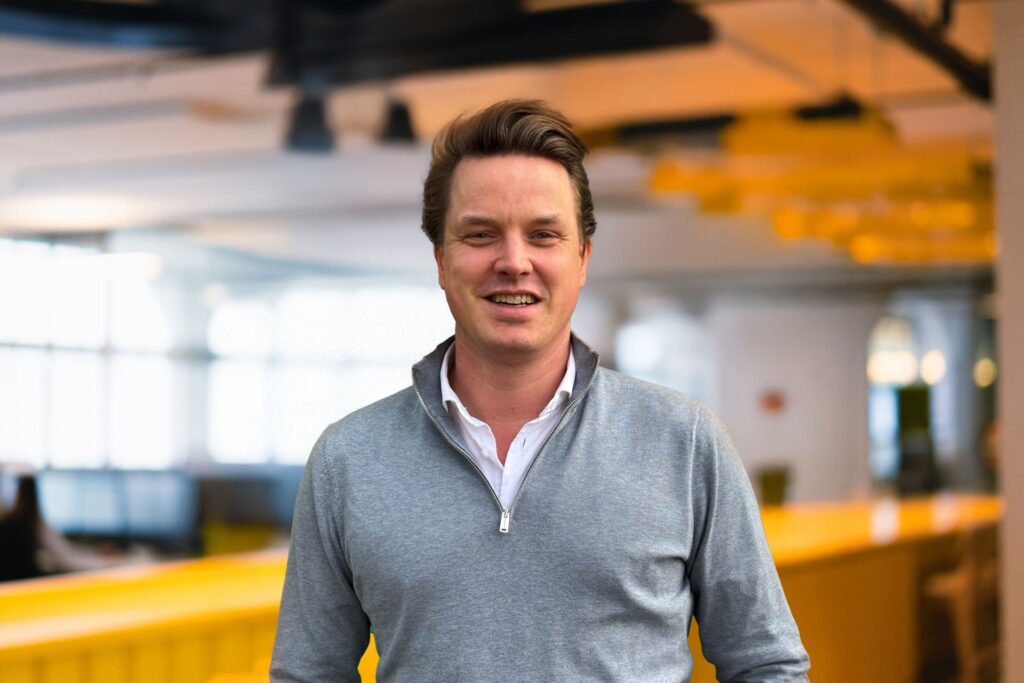 Man met kort bruin haar en grijze trui lacht in een moderne kantoorruimte met gele accenten op de achtergrond.